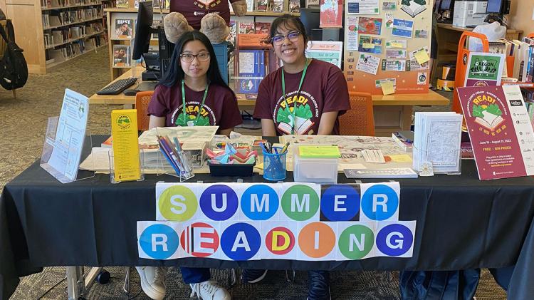 Two teen volunteers seated at the Summer Reading table inside the library
